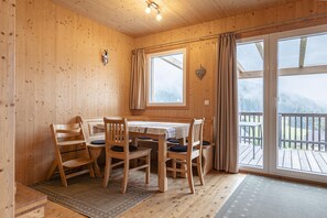 Ein Holzinterieur mit einem Esstisch und Stühlen, einem Fenster mit Vorhängen und Blick auf eine Terrasse.