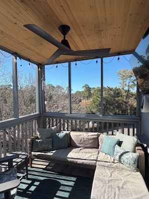 A covered outdoor seating area with a ceiling fan, string lights, and a view of trees.