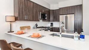 A modern kitchen with dark wood cabinets, a stainless steel refrigerator, a microwave, and a gas stove. There are two orange bowls on the counter, a lamp, and a sign with food items.