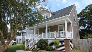 A two-story house with a covered front porch, a small front yard, and a wooden fence. - Buckroe Beach Home with King Bed, Grill, Games and 5 min Walk to Beach…Sleeps 8. (Hampton)