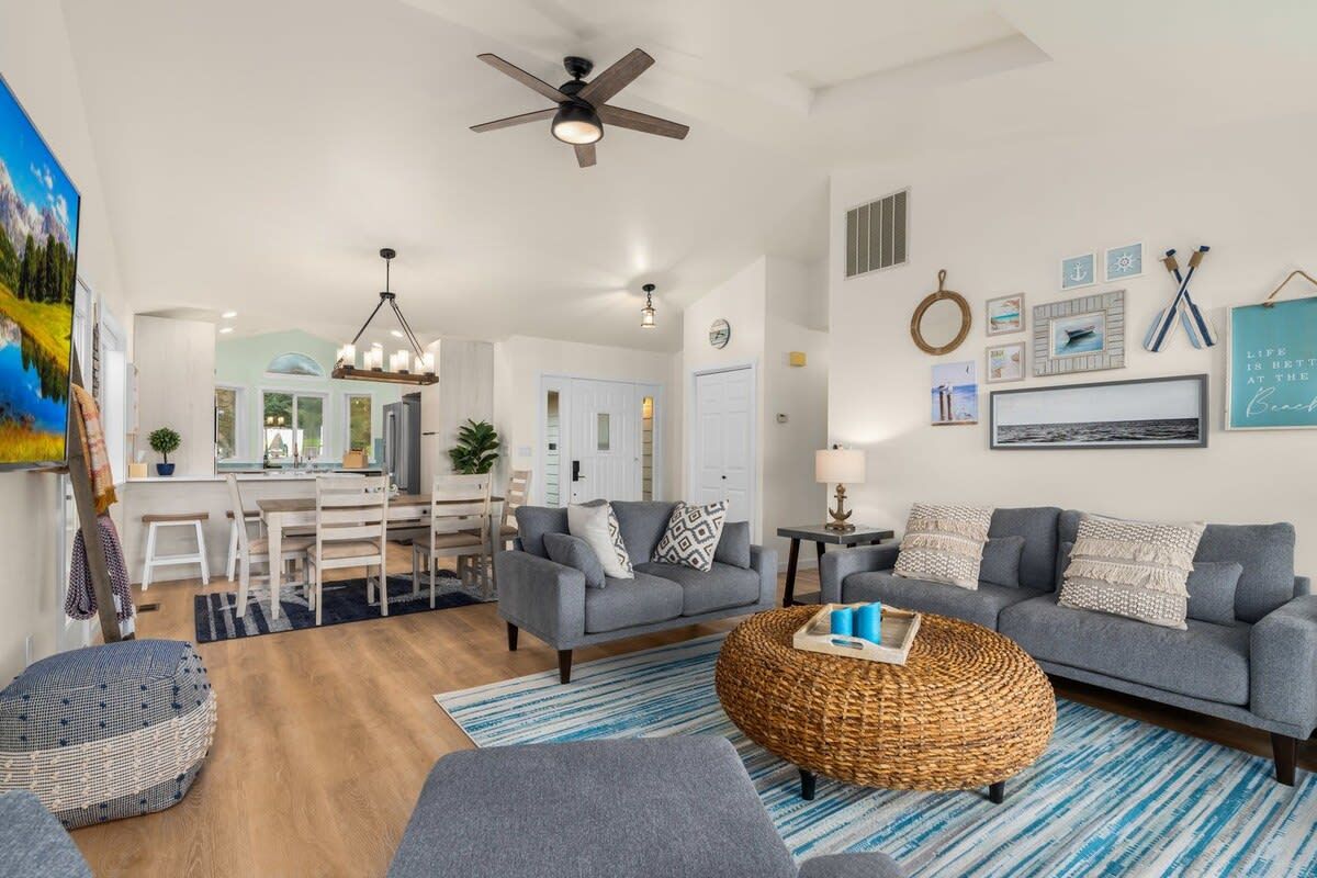 A spacious living room with a grey sofa, a round wicker coffee table, and a blue-striped rug. There is a dining area with a white table and chairs, and a kitchen in the background.