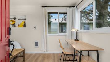 A room with a wooden table and chair, a red door, and a window with white curtains.