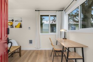 A room with a wooden table and chair, a red door, and a window with white curtains. - @ Marbella Lane – Wave HS w/EV Charger, Oceanfront! (Oak Harbor)