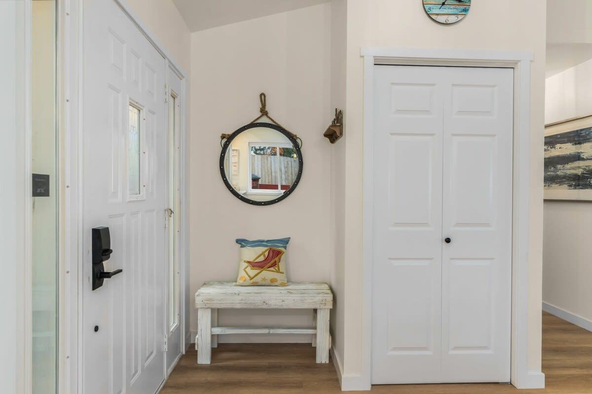 Un couloir avec une porte blanche, un banc doté d’un coussin, un miroir fixé au mur et une horloge.