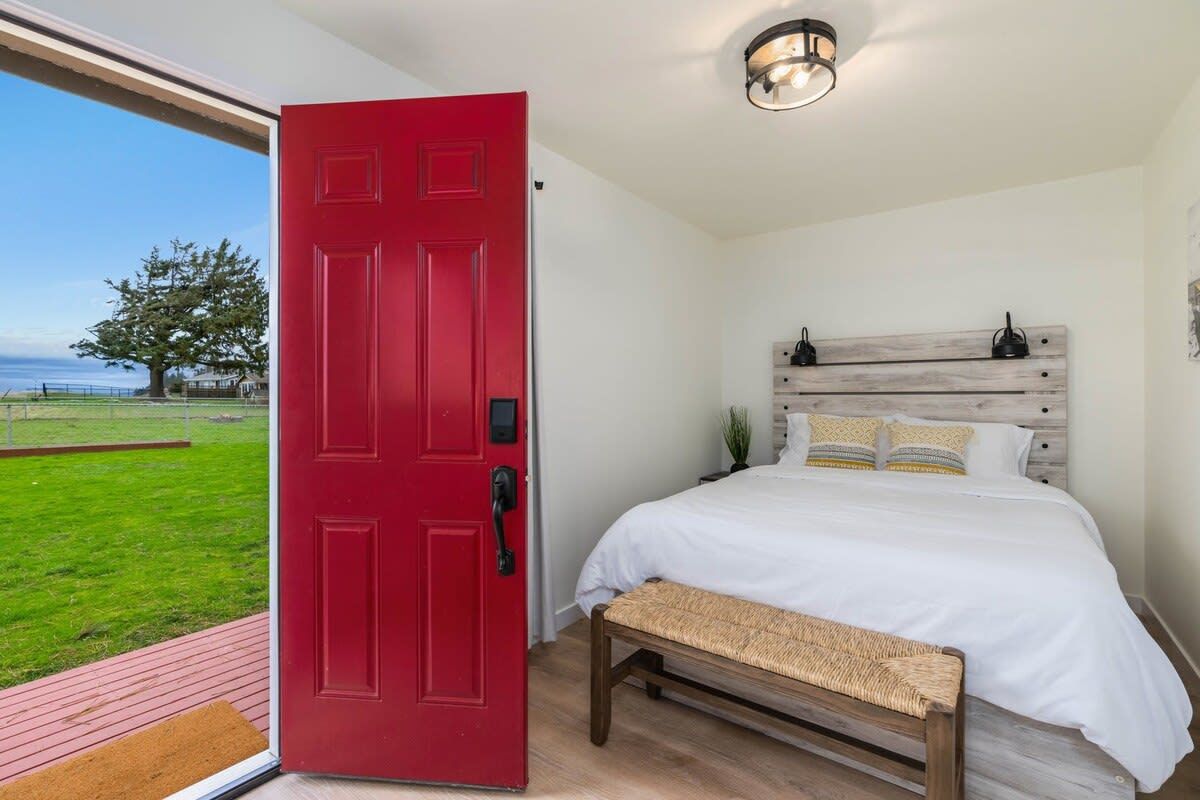 A bedroom with a red door, a bed with white bedding, a wooden headboard, and a view of a grassy area and a tree.