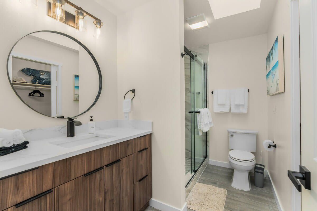 A modern bathroom with a wooden vanity, a large mirror, a glass shower enclosure, and a toilet.