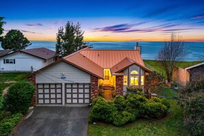 A house with a garage, a driveway, and a view of the ocean at sunset. - @ Marbella Lane – Wave HS w/EV Charger, Oceanfront! (Oak Harbor)