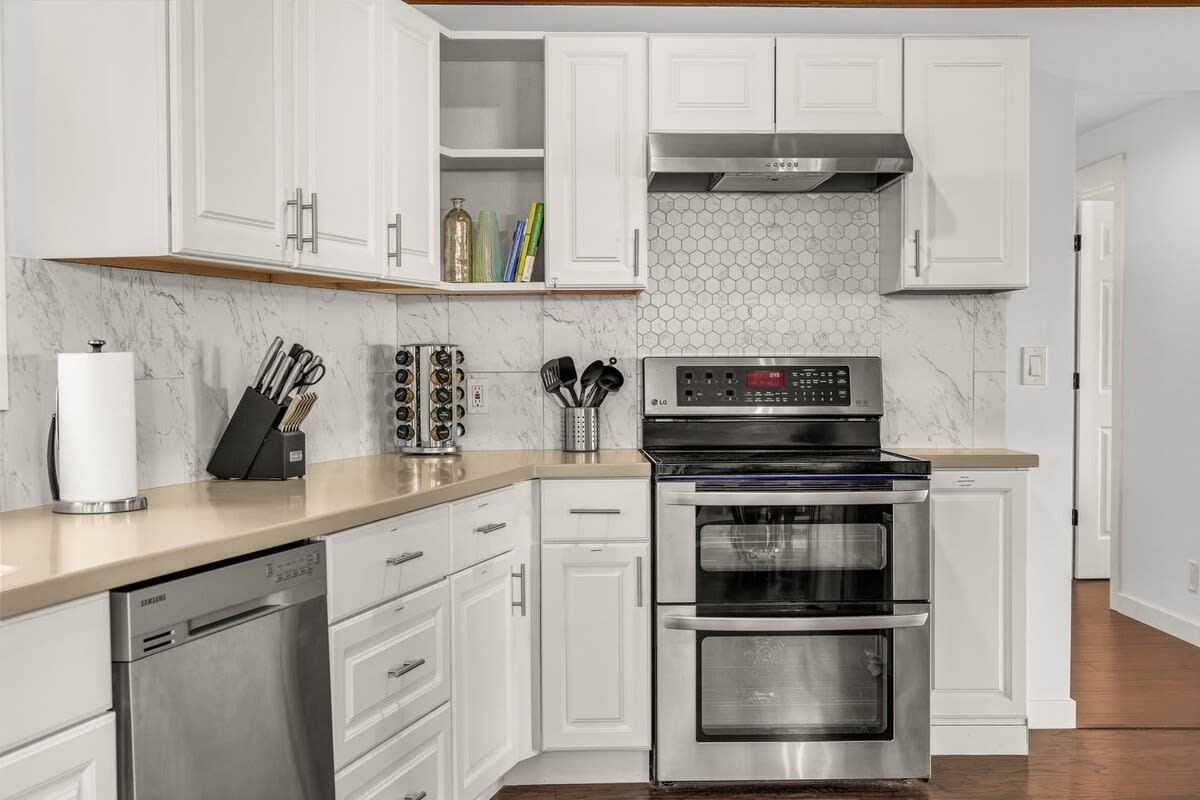A modern kitchen with white cabinets, a stainless steel dishwasher, a double oven, and a hexagonal tile backsplash.