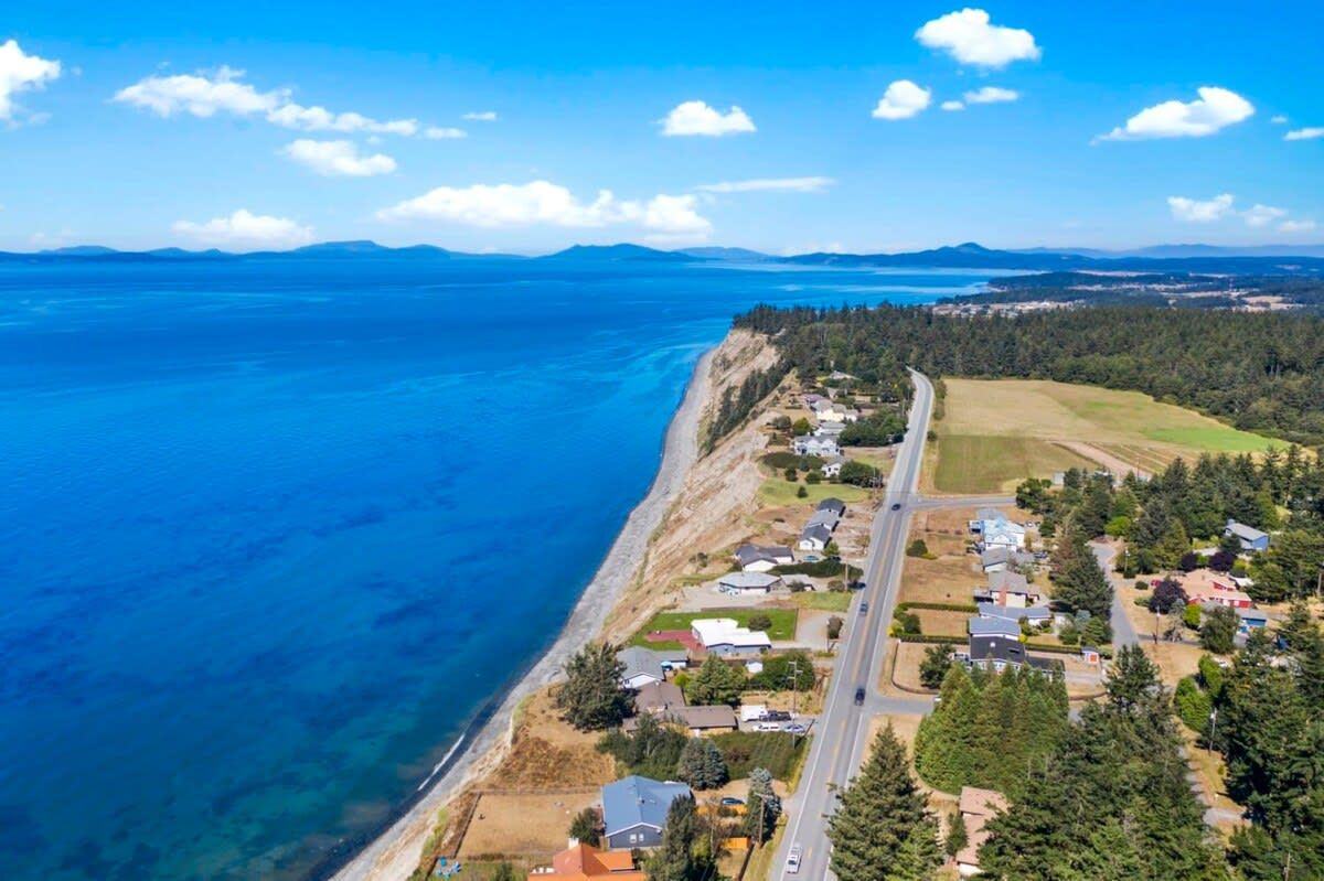 An aerial view of a coastal residential area with houses along the shoreline, a road, and a clear blue sea.