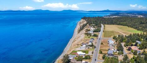 An aerial view of a coastal residential area with houses along the shoreline, a road, and a clear blue sea.