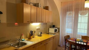 Ett kompakt kök med träskåp, en mikrovågsugn, en vattenkokare och ett handfat. Här finns ett matbord med stolar och ett fönster med tunna, genomskinliga gardiner.