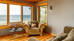 Une pièce chaleureuse avec un plafond en bois, de grandes fenêtres donnant sur la plage, un fauteuil confortable et une petite table agrémentée d’un objet décoratif.