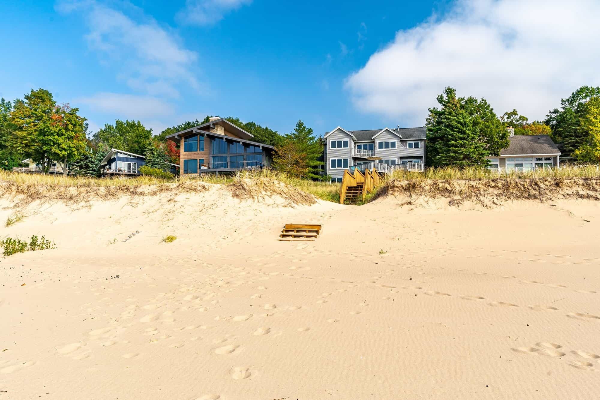Une propriété située en bord de mer, dotée d’une plage de sable, d’un escalier en bois menant à une terrasse en bois, et de plusieurs maisons avec de grandes fenêtres.