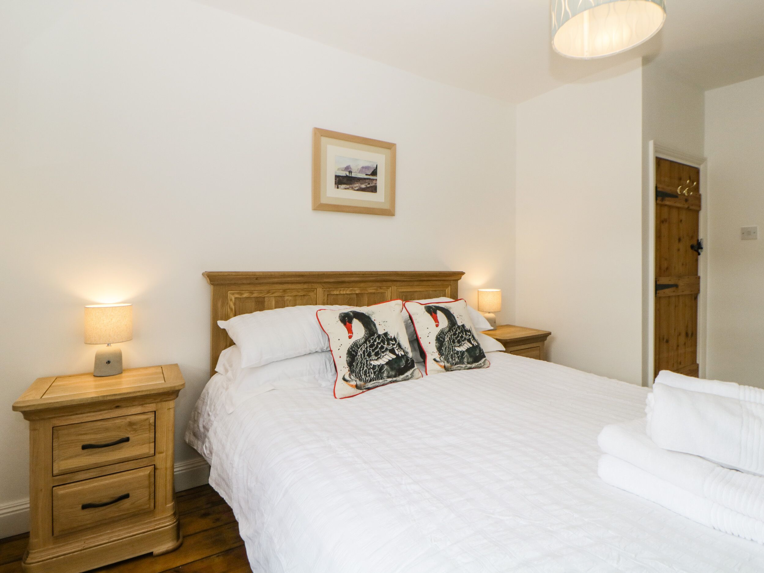 Cama bem arrumada com roupa de cama branca e duas almofadas decorativas com cisnes pretos. Há duas mesas de cabeceira com abajures, uma cabeceira de madeira e um quadro na parede.
