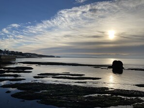A coastal landscape with a rocky shoreline, calm water, and a distant sun setting or rising. - The Yellow House, 13 King Street (Dawlish)