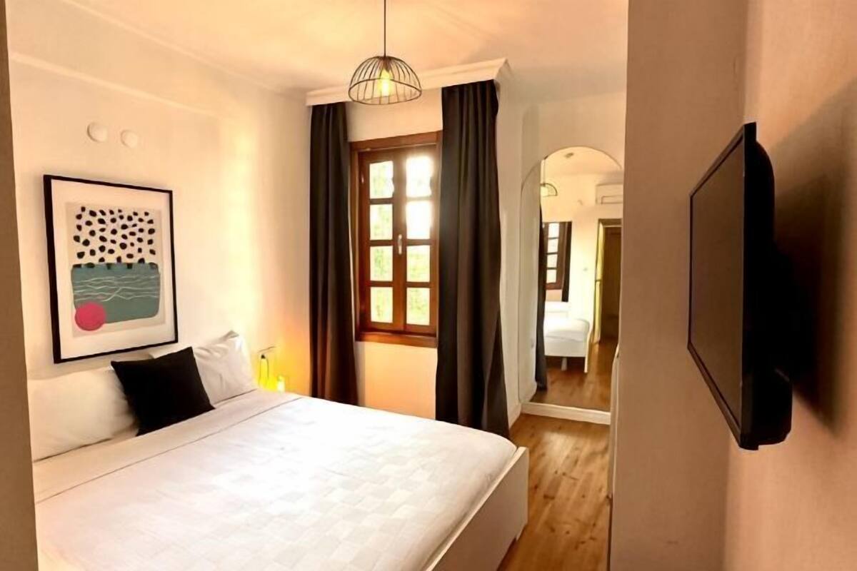 A bedroom with a bed, a wall-mounted TV, a framed picture, and a window with curtains.