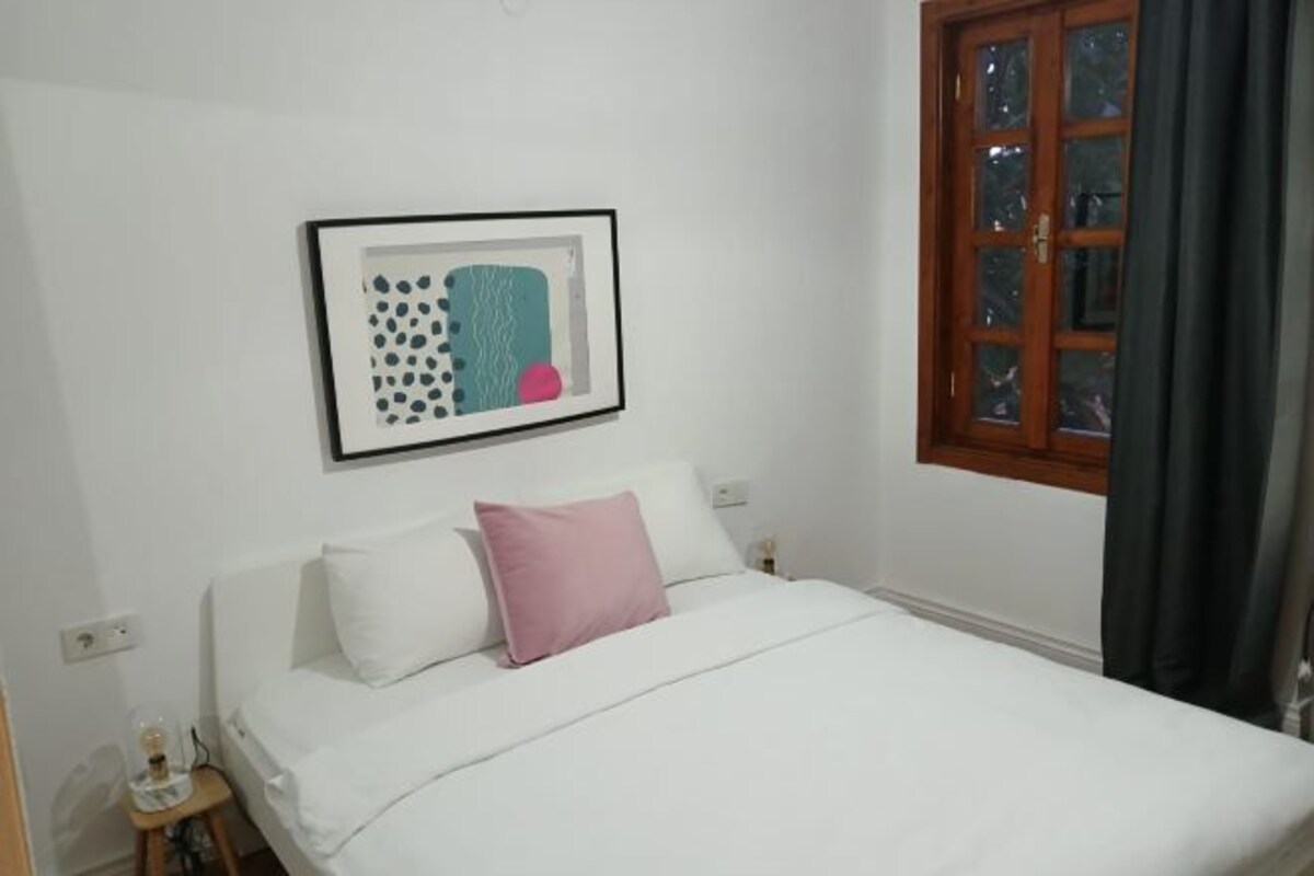 A bedroom with a bed, a nightstand, a framed picture, and a window with curtains.