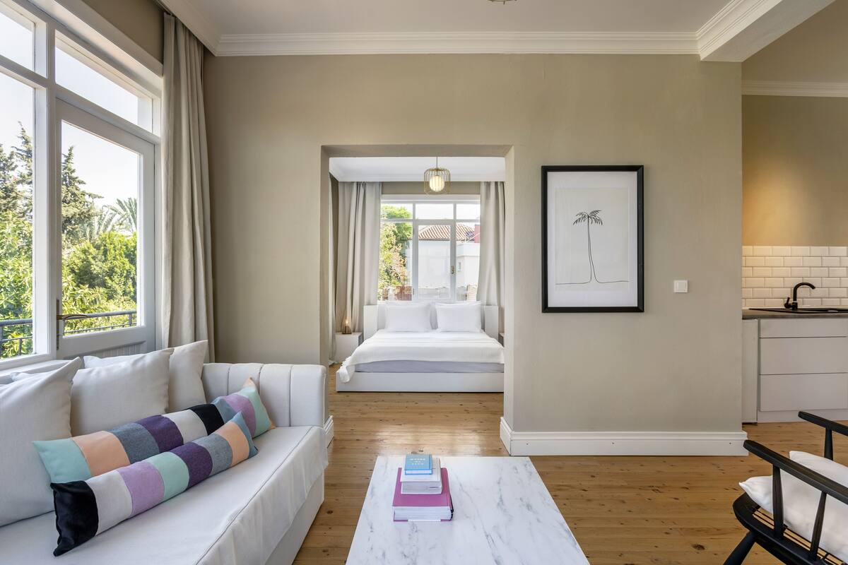 A modern hotel room with a large bed, a sofa, a marble coffee table with books, a framed picture on the wall, and a view of the outdoors through the window.