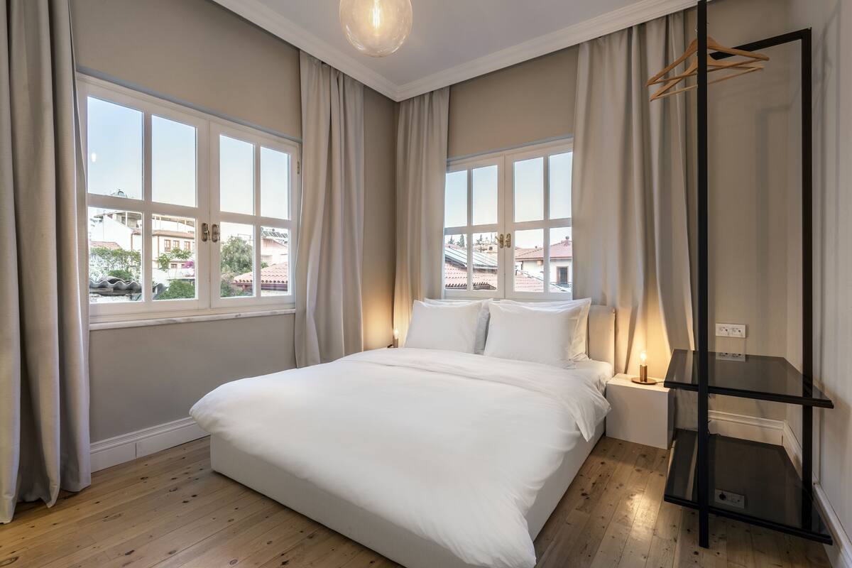 A bedroom with a large bed, two bedside tables, a black shelf unit, and a view of buildings through large windows.