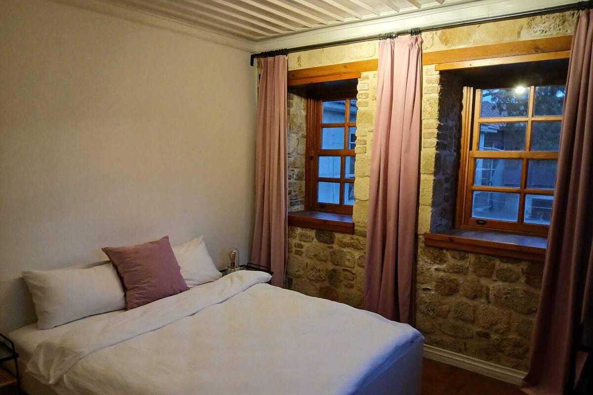 A bedroom with a bed, two windows with curtains, stone walls, and a wooden beam ceiling.