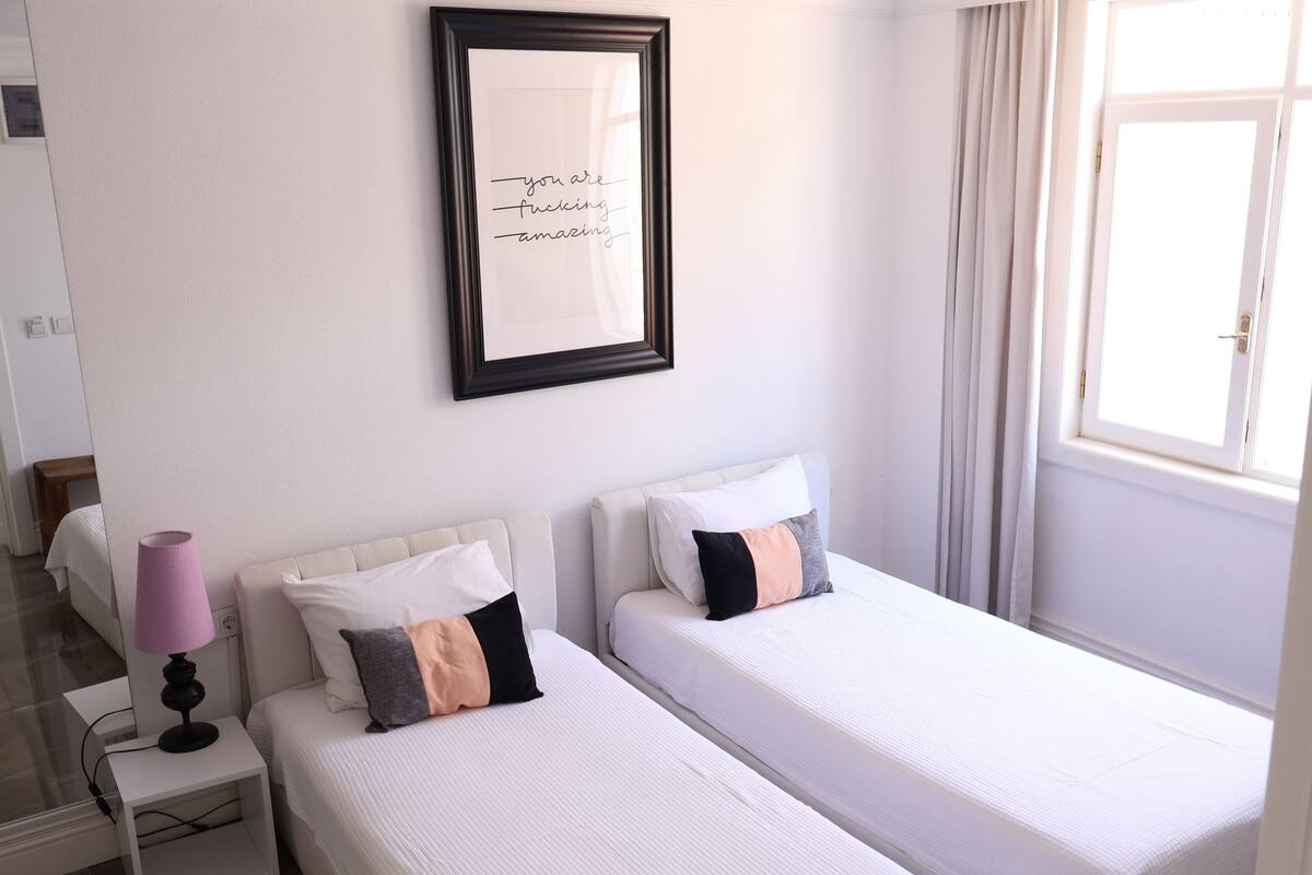 A hotel room with two beds, a bedside table with a lamp, a framed quote on the wall, and a window with curtains.