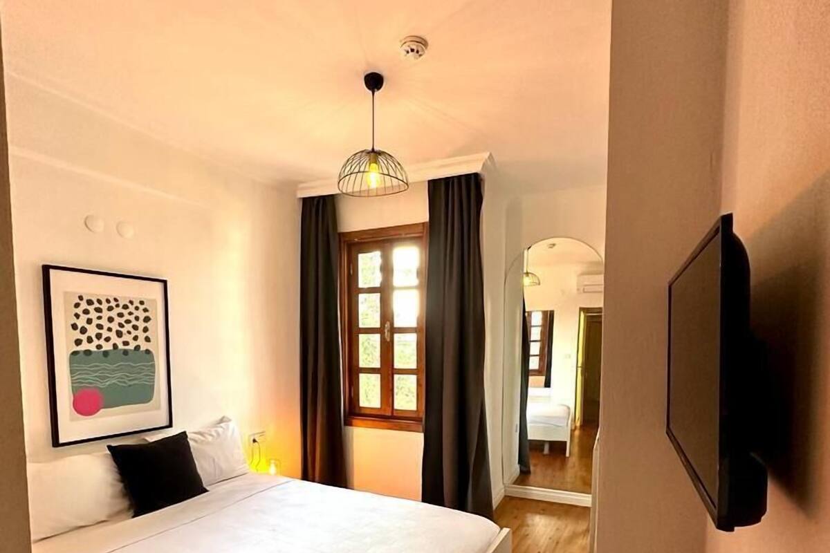 A hotel room with a bed, a flat-screen TV mounted on the wall, a framed picture on the wall, and a window with curtains.