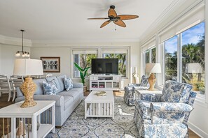 A spacious living room with a large TV, white furniture, and a ceiling fan.