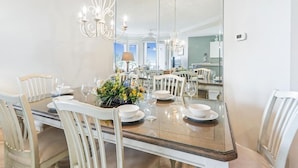 Un coin repas agrémenté d’un grand miroir, de chaises blanches, d’une table en verre et d’un lustre.