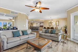 Un salon spacieux avec deux canapés, une table basse, un ventilateur de plafond et une cuisine en arrière-plan.