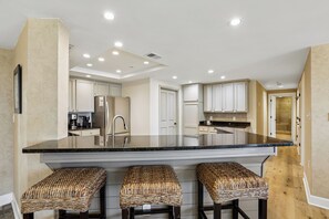 A kitchen with a central island, three wicker bar stools, white cabinets, and a stainless steel refrigerator. - Ocean Front 4Br Villa w/ Large Balcony! (Hilton Head Island)