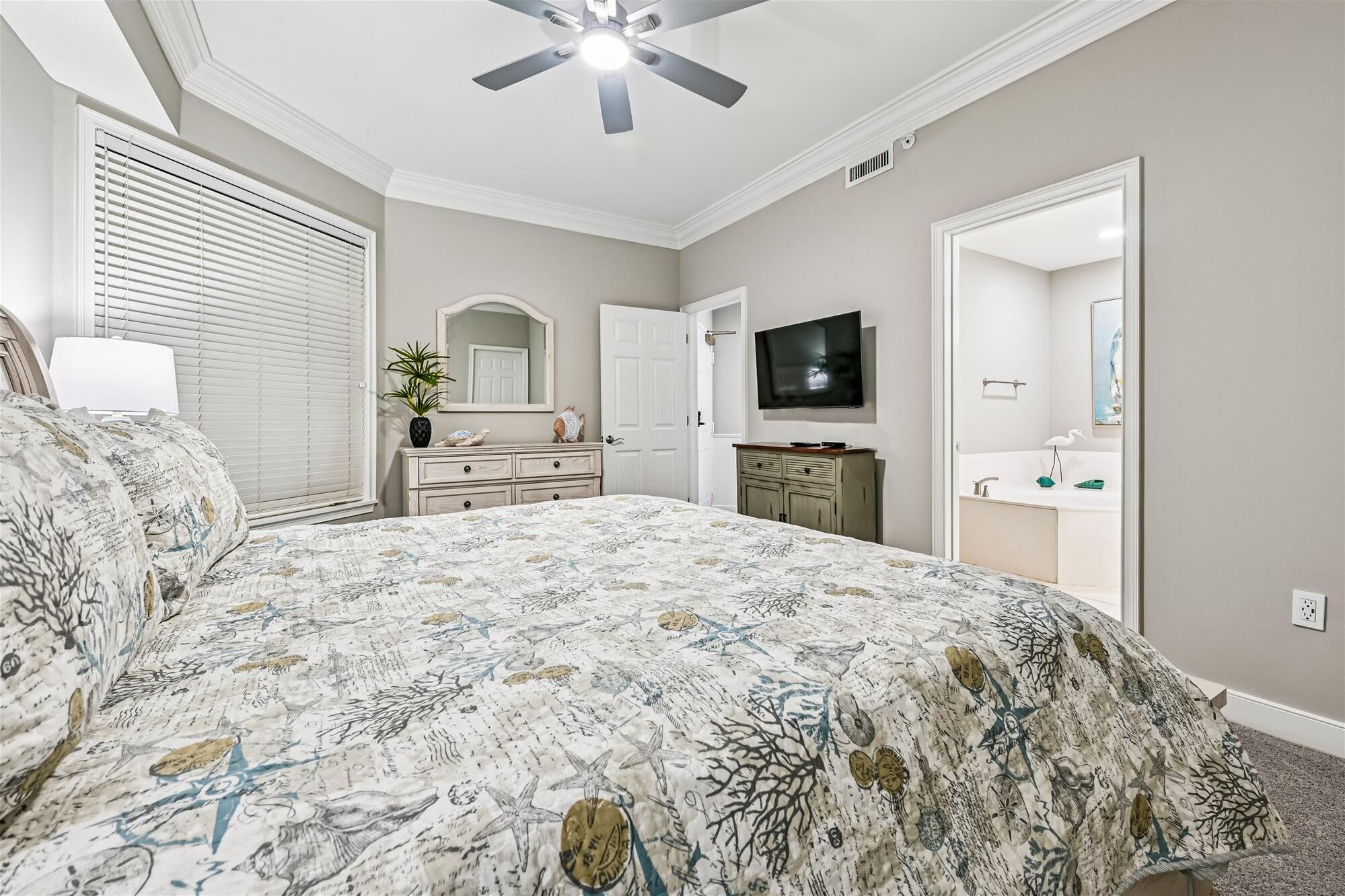 A bedroom with a large bed, a ceiling fan, a television, a dresser, and a bathroom with a sink and mirror.