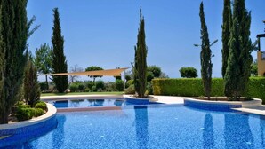 A swimming pool with a clear blue water feature, surrounded by neatly trimmed hedges and tall cypress trees. - Apartment A02 - Roudias (Kouklia)