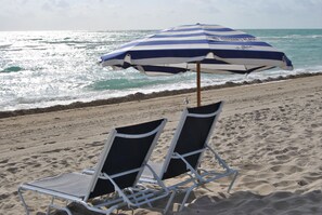 Ein Strand mit zwei weißen Liegestühlen und einem blau-weiß gestreiften Sonnenschirm.