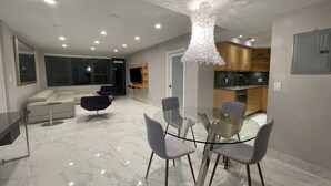 A modern living room with a glass dining table, grey chairs, a white sofa, and a large TV on a wooden stand. - The Alexander Resort Ocean Front (Miami Beach)