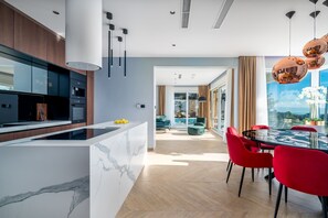 A modern kitchen with a marble island, wooden cabinets, and a dining area with a glass table and red chairs.