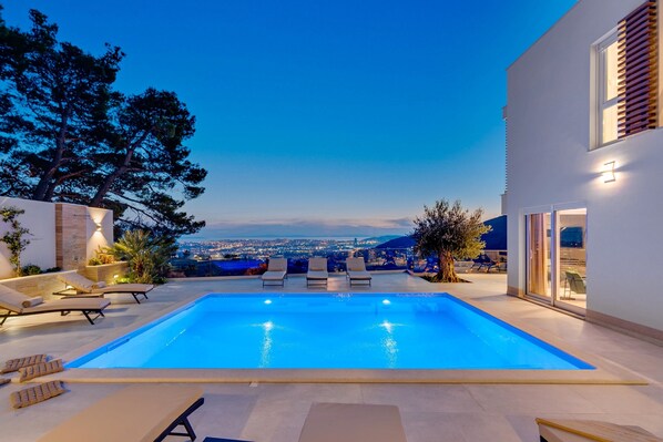 A modern pool area with lounge chairs and a view of the city at dusk. - Luxury Villa Seventh Heaven with heated pool, hot tub, gym, panoramic views (Klis)