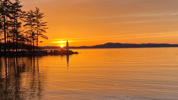 A serene sunset over a calm lake with a lighthouse and trees on the shore. - 6 bedroom waterfront cabin -enjoy beautiful sunsets! (Frye Island)