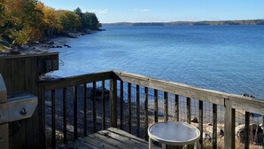 A wooden deck with a grill and a table overlooking a calm body of water. - 6 bedroom waterfront cabin -enjoy beautiful sunsets! (Frye Island)