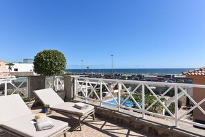 Tagterrasse med liggestole, et lille bord med hat og håndklæde, samt udsigt over havet og parkeringsområde.