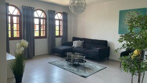 Ein modernes Wohnzimmer mit einem dunklen Sofa, einem Glastisch und einem großen Fenster mit Vorhängen.