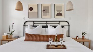 Een net opgemaakt bed met een bruin kussen, een dienblad met twee wijnglazen en een fles wijn. Drie aan de muur bevestigde fotolijstjes en twee hangende rieten lampen.