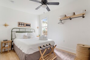 Une chambre à coucher comprenant un lit, un ventilateur de plafond, une fenêtre, une petite table, une étagère et une corbeille à déchets.