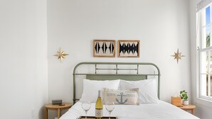 A neatly made bed with white linens, a tray with a bottle and glasses, and a pillow with an anchor design. - Only 2 Blocks to King! | The Murray (Charleston)