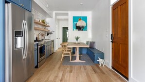 A modern kitchen with a built-in refrigerator, oven, and sink. There is a dining area with a table and chairs, and a wooden bench. A small dog is visible near the door. - Only 2 Blocks to King! | The Murray (Charleston)