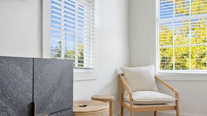 Une pièce avec un buffet en bois foncé, un tabouret en osier, une chaise en bois avec un coussin et une fenêtre avec des stores blancs.