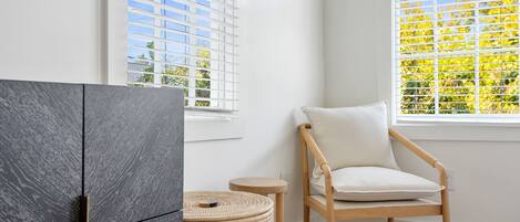 Una habitación con un armario de madera oscura, un taburete de mimbre, una silla de madera con cojín y una ventana con persianas blancas.