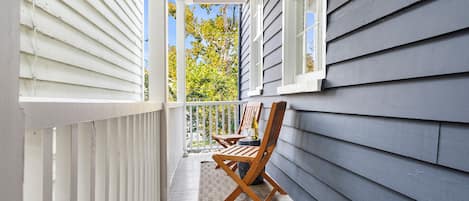 A covered porch with a wooden chair and a small table, white railings, and a view of trees and sky.