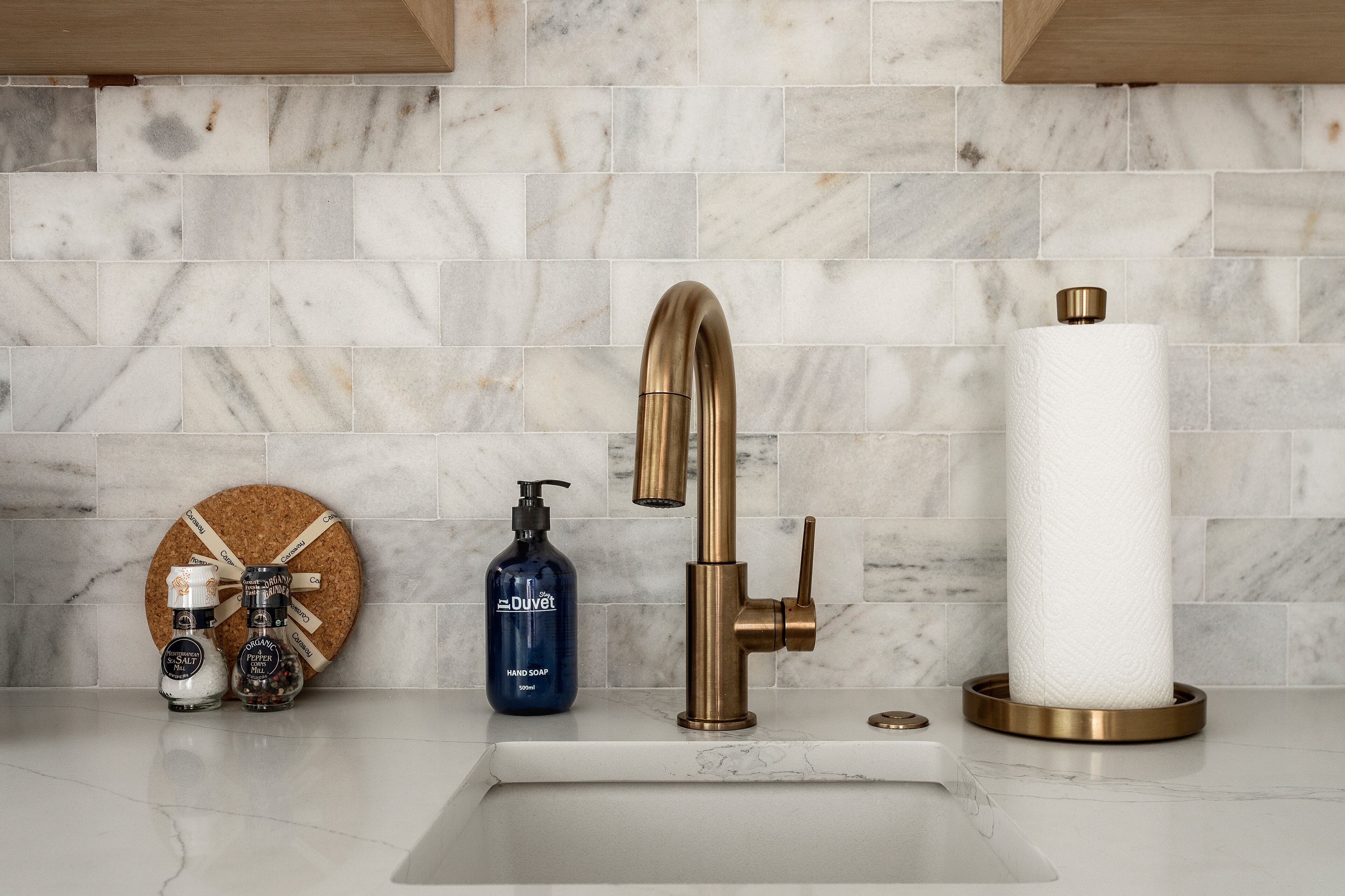 A bathroom with a marble backsplash, a gold faucet, a soap dispenser, and a paper towel holder.