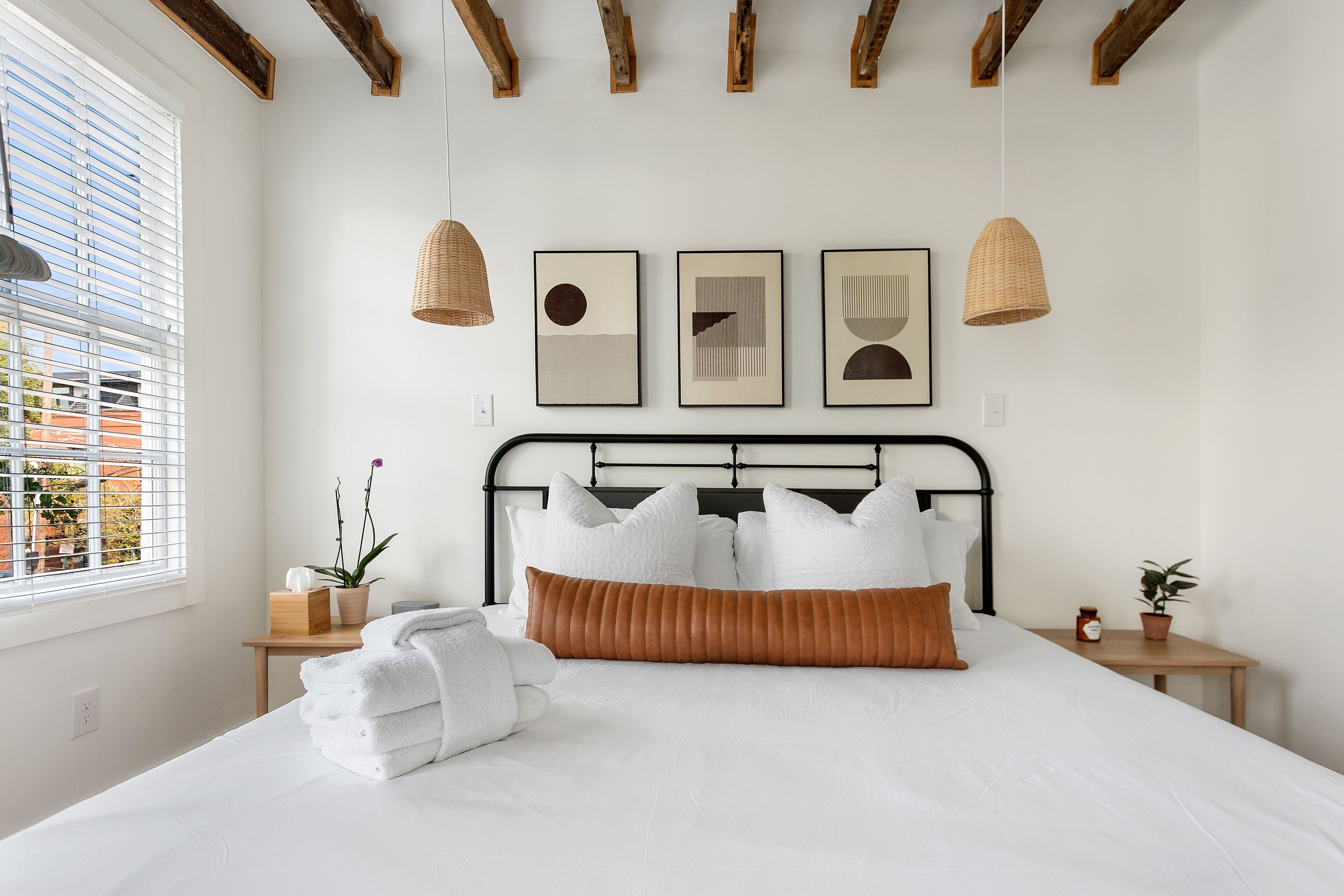 A neatly made bed with white linens and a brown accent pillow, a bedside table with a plant, and a window with blinds.