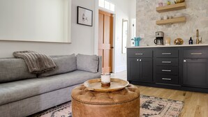 Un salon avec un canapé gris, un pouf marron et une table basse. On y trouve également un coin cuisine avec des meubles sombres et un plan de travail.